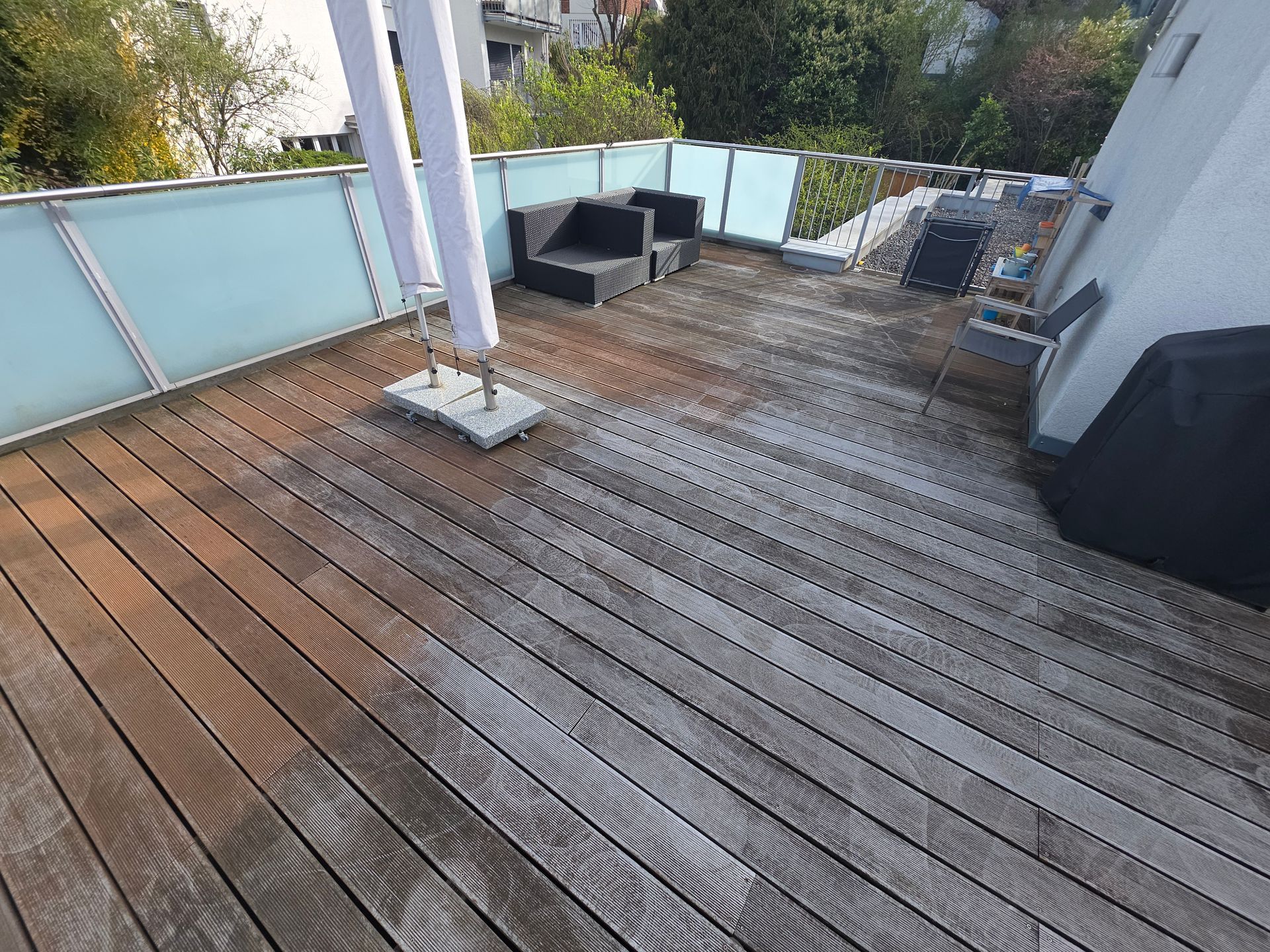 Eine hölzerne Dachterrasse mit Terrassenmöbeln, Glasgeländer und einem kleinen Tisch in der Nähe eines weißen Gebäudes.