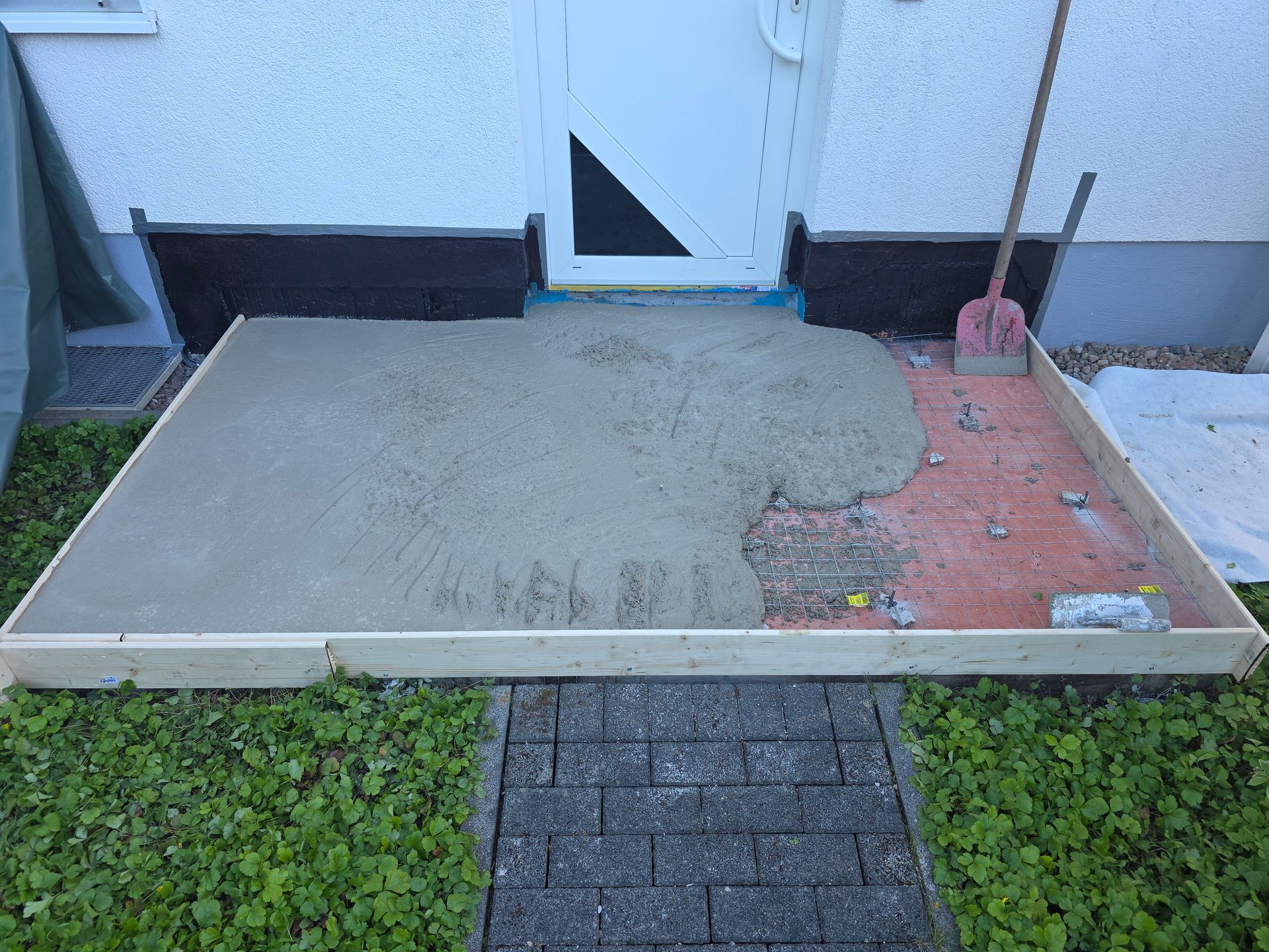 Kleine Betonveranda mit einer Reparaturstelle aus roten Ziegelsteinen, vor einer Tür und einer Mauer.