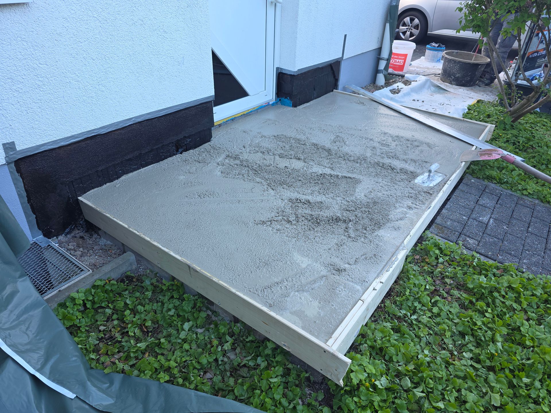 Betonplatte neben einem weißen Gebäude, im Vordergrund eine schwarze Stützstrebe und Gras.