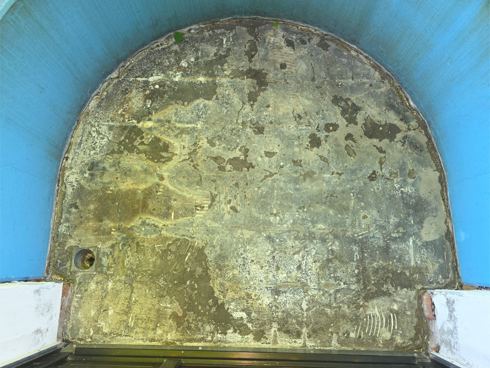 Verwitterte Steinplatte mit abgerundetem Ende, eingefasst zwischen blauen Mauern, mit einem kleinen Loch in der Nähe der oberen linken Ecke.