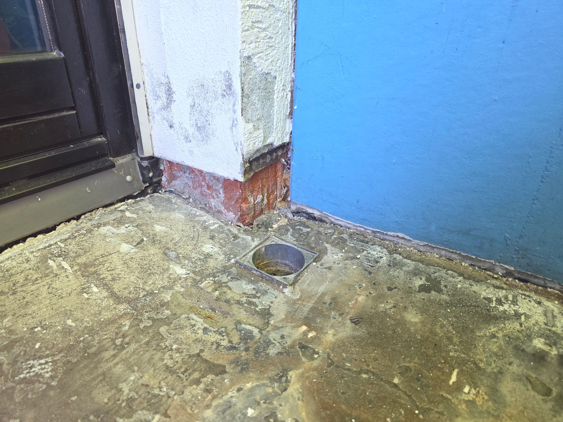 Beschädigter Betonboden mit einem Abfluss an der Ecke eines Gebäudes, neben einer blauen Wand und einer Tür.