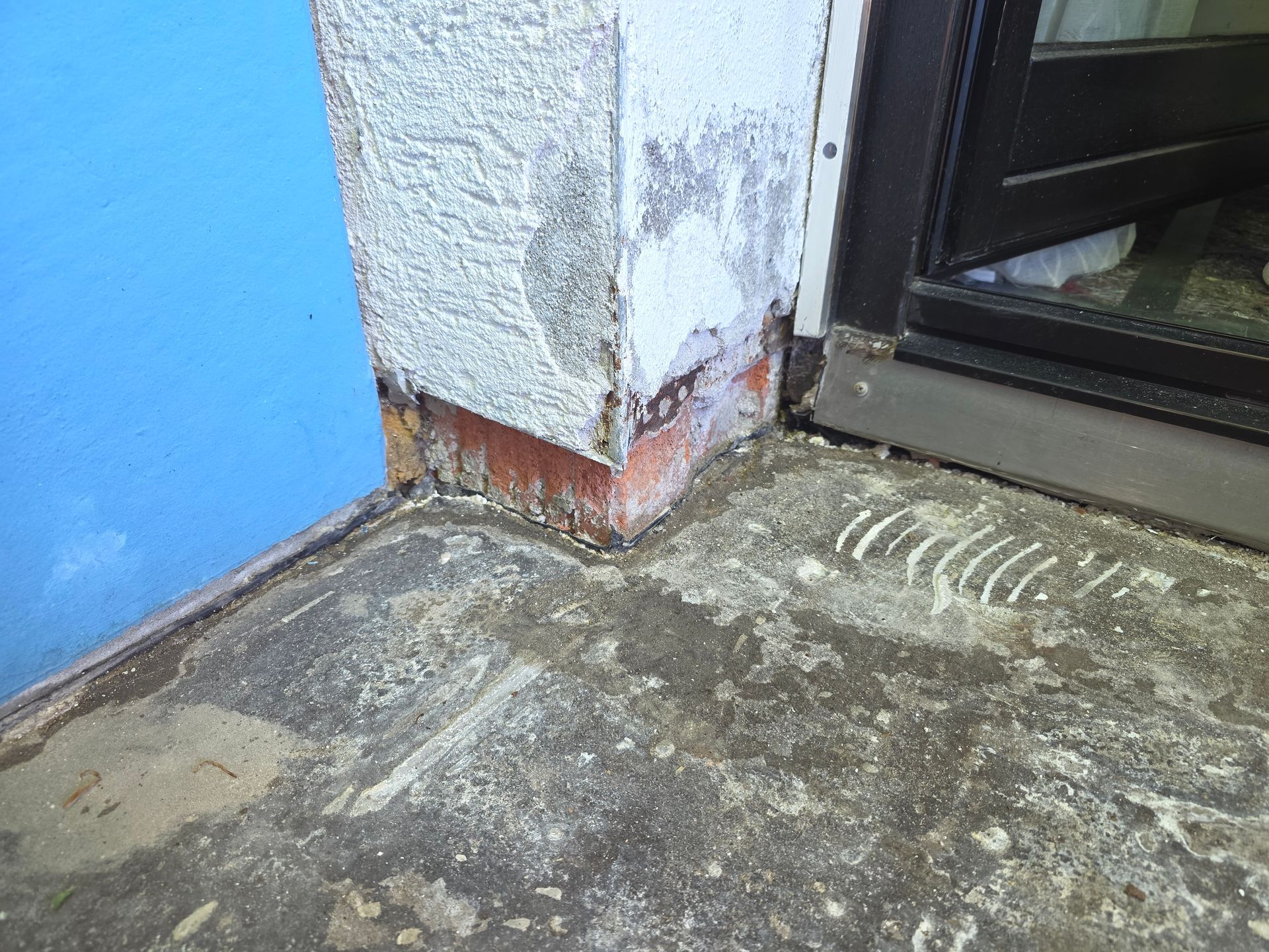 Beschädigte Außenwandecke mit abblätterndem Putz und rissigem Beton neben einer Türschwelle