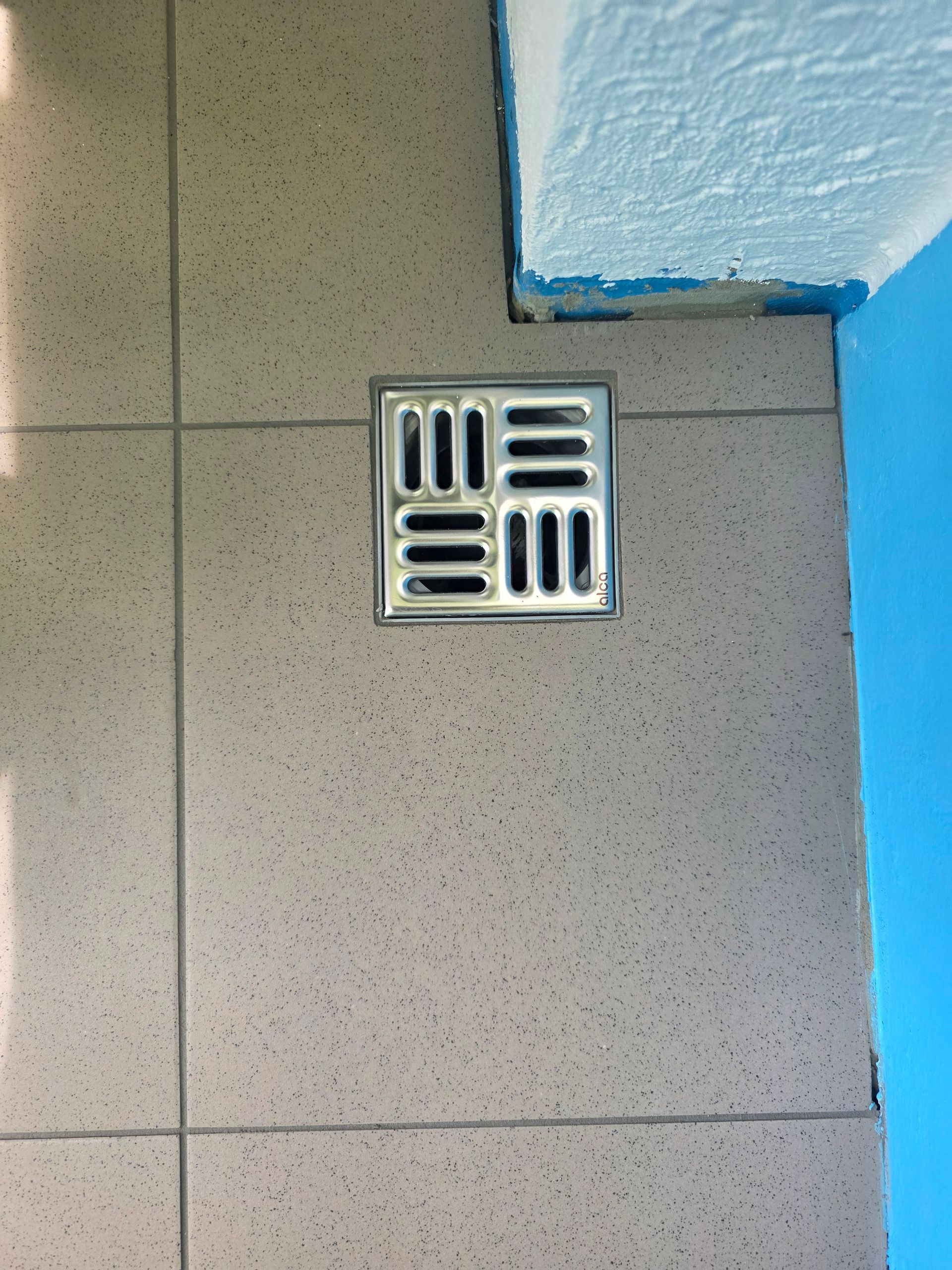 Quadratischer Metall-Bodenablauf auf beigefarbenem Fliesenboden neben einer blauen Wandecke.