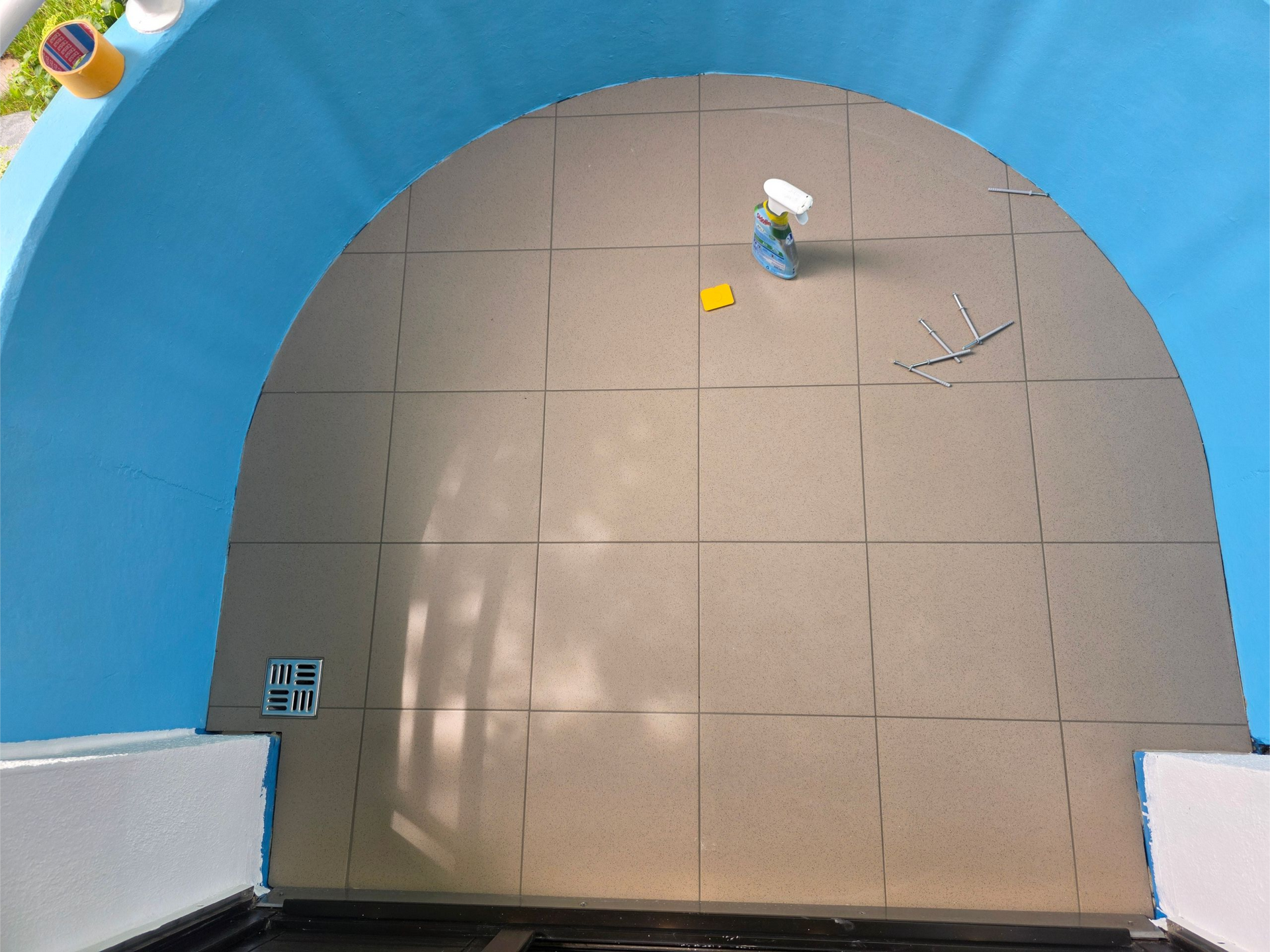 Ein leerer Fliesenboden neben einer geschwungenen blauen Wand, in der Ecke stehen ein Wischmopp und ein Eimer.