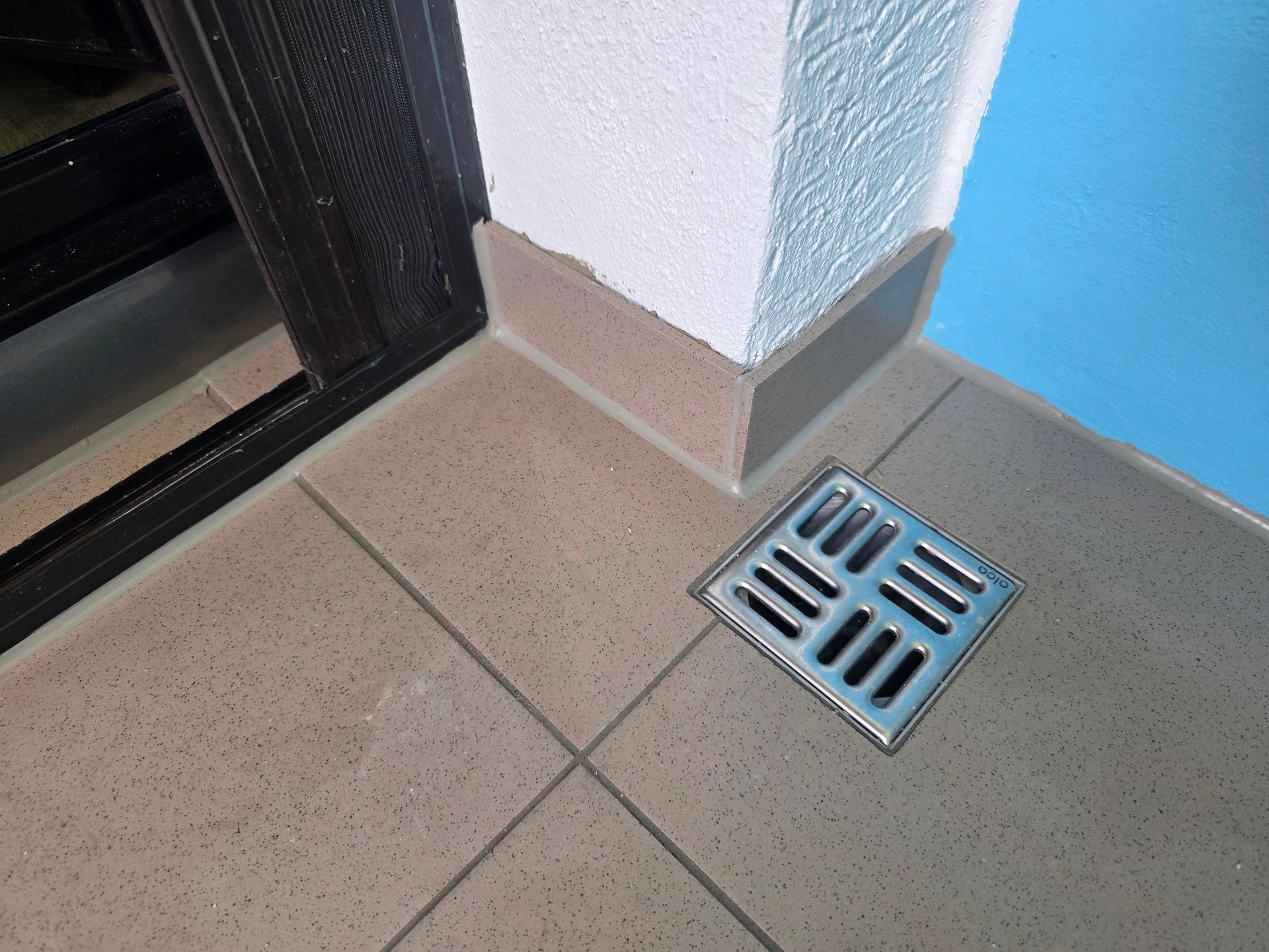 Bodenablaufgitter in einer beige gefliesten Ecke neben einer weißen Wand und einer blauen Oberfläche