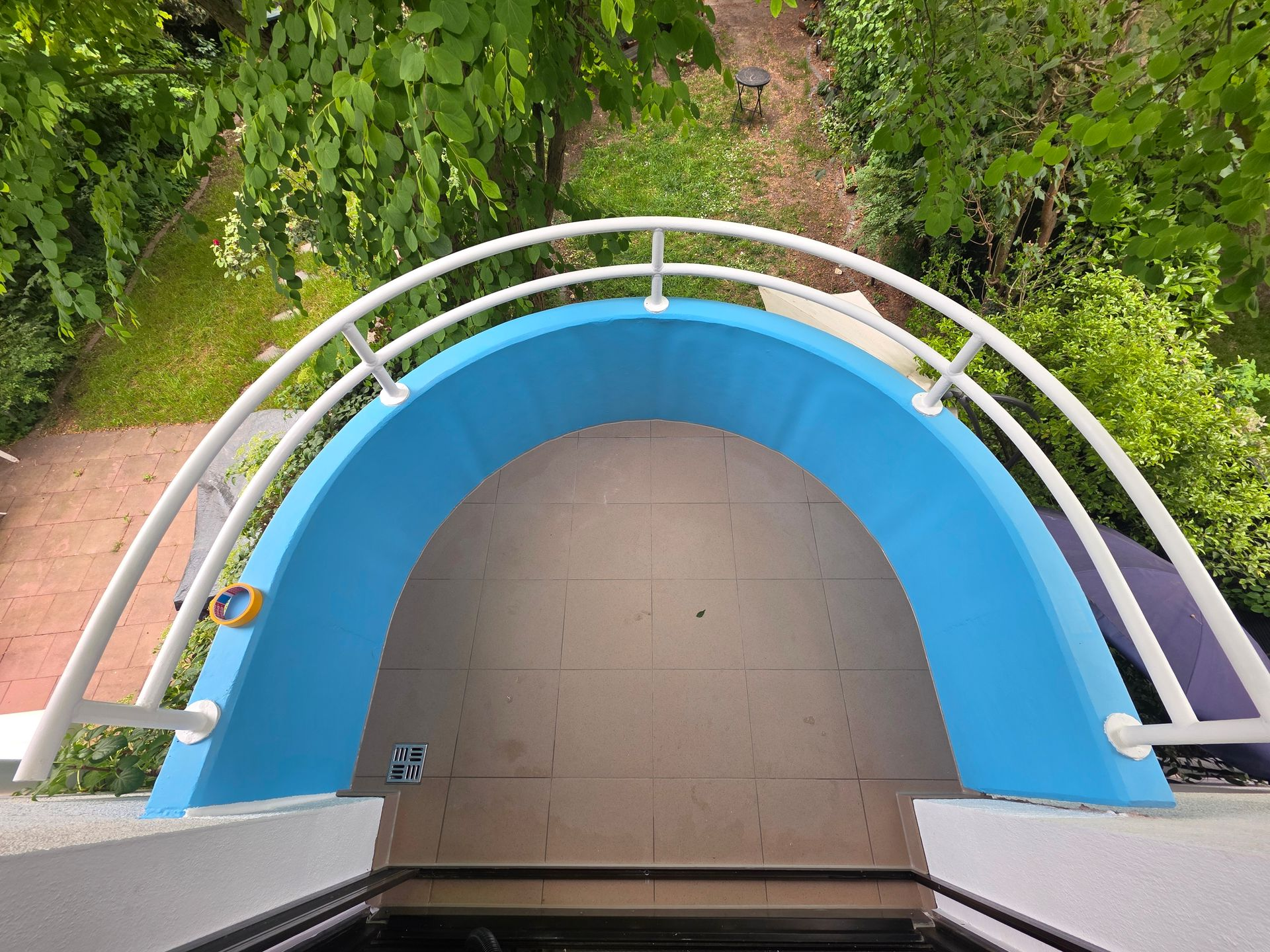 Blaues Trampolin im Garten, von oben gesehen, mit weißen Geländern und grünen Pflanzen drumherum