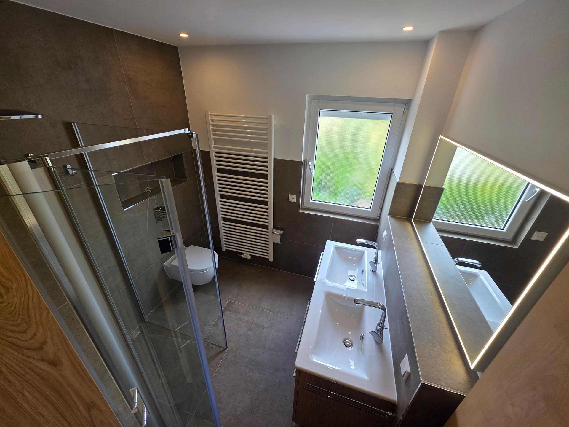 Modernes Badezimmer mit Glasdusche, weißem Waschbecken, Handtuchhalter und zwei Fenstern mit Jalousien.