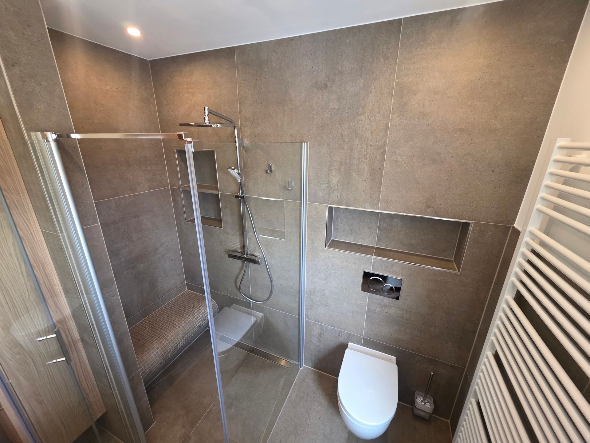 Modernes Badezimmer mit Glasdusche, WC, grau gefliesten Wänden und weißem Handtuchheizkörper.
