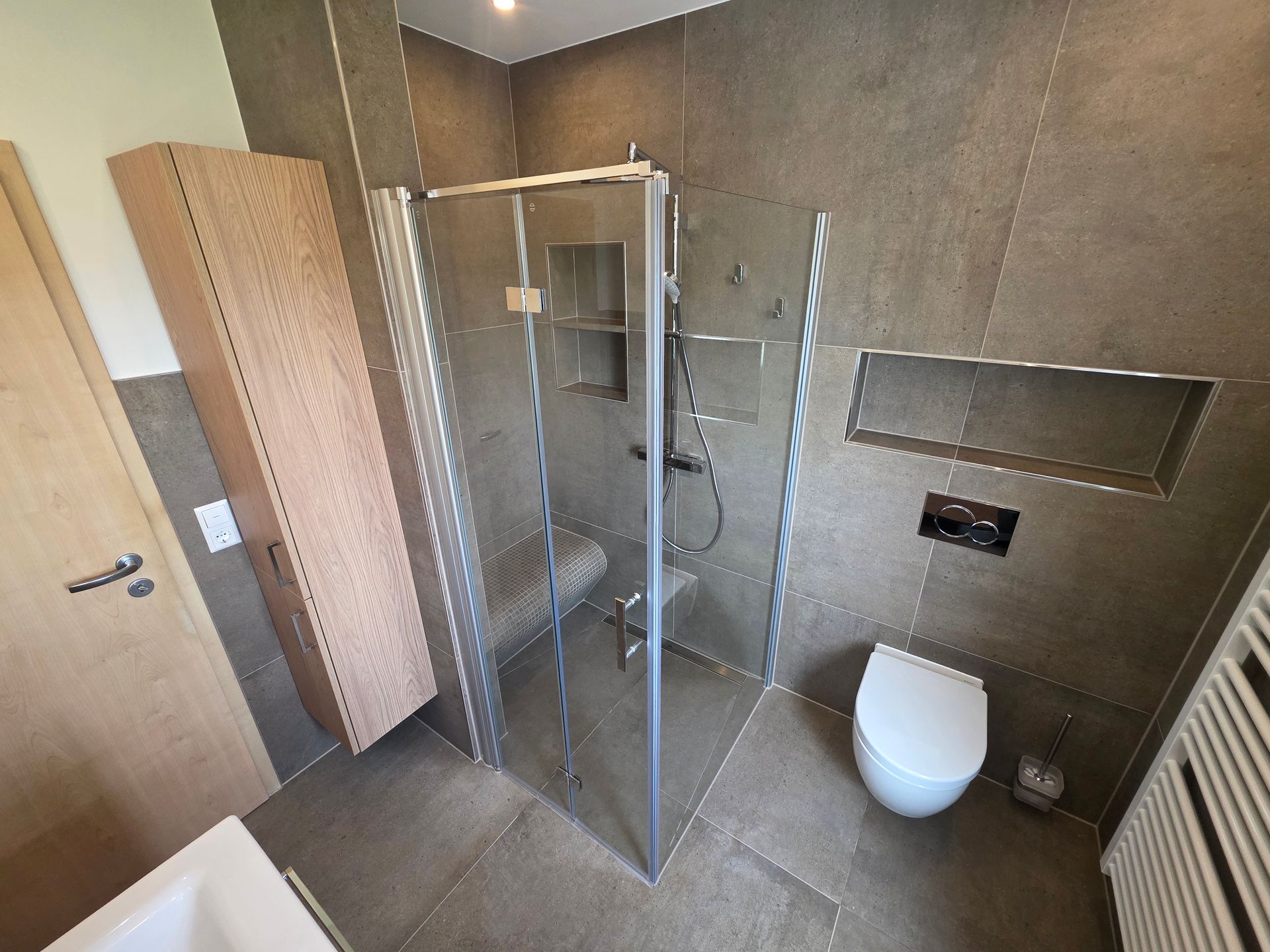 Modernes Badezimmer mit ebenerdiger Glasdusche, weißer Toilette, grau gefliesten Wänden und hohem Schrank.