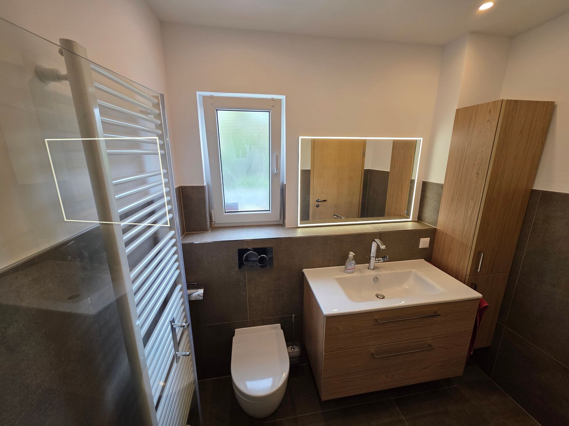 Modernes Badezimmer mit Toilette, Waschtischunterschrank, Spiegel, Handtuchheizkörper und Fenster mit Milchglas.