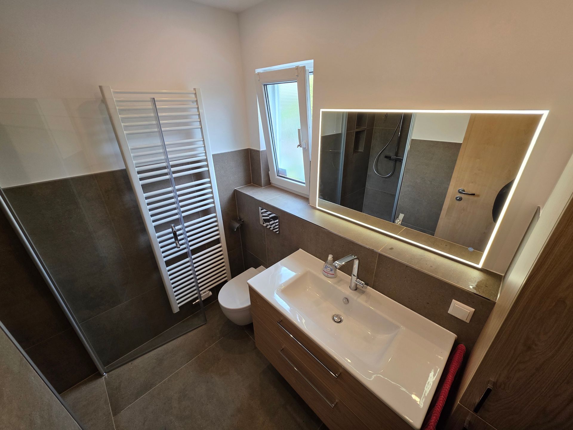 Modernes Badezimmer mit weißem Waschbecken, Spiegel, Handtuchheizkörper, Toilette und dunklen Fliesenwänden