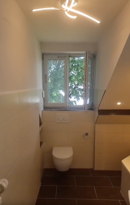 Kleines Badezimmer mit Toilette unter einem Fenster und moderner Deckenleuchte.