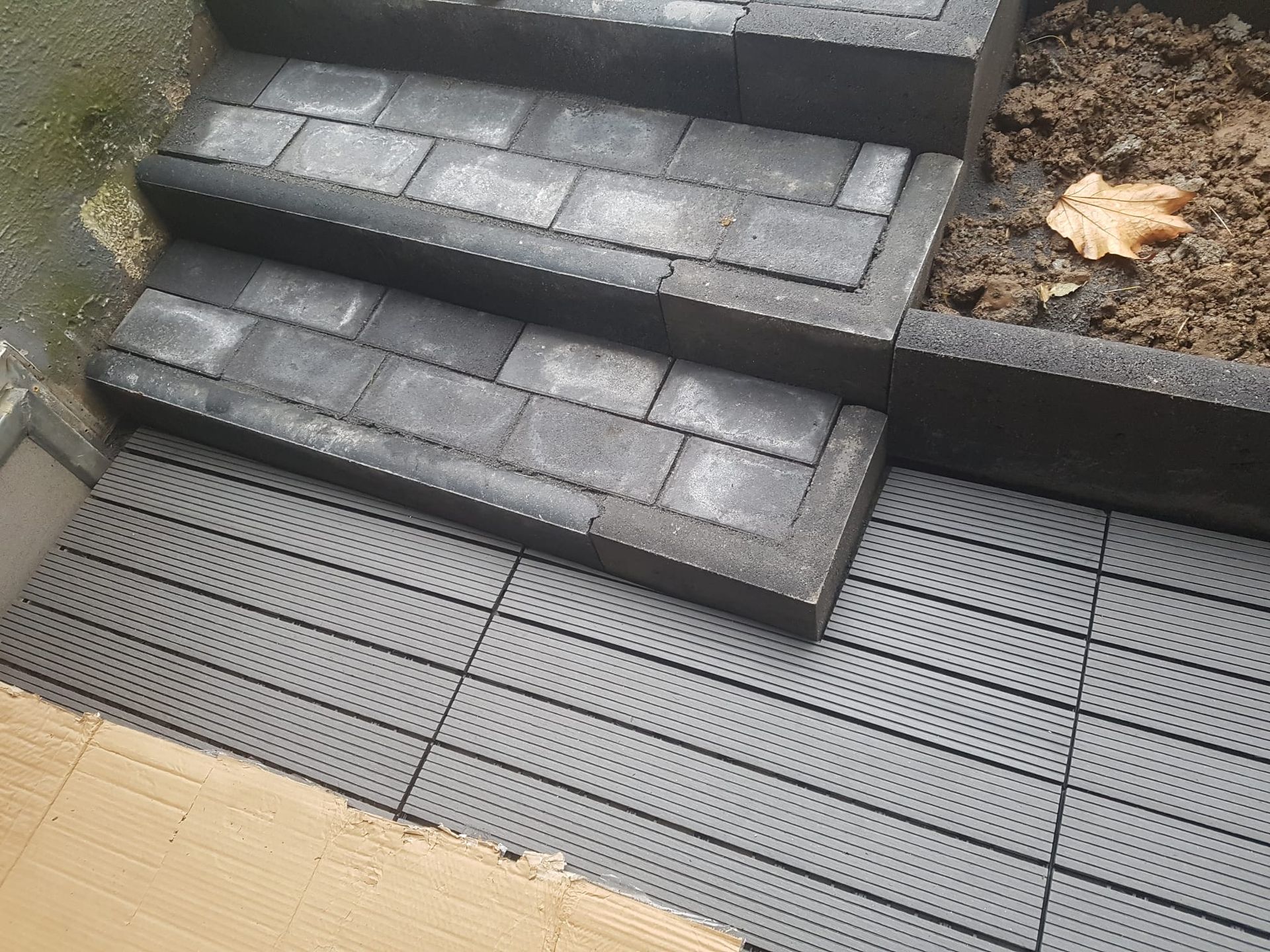 Außentreppe mit dunklen Pflastersteinen und ein grauer Gitterweg neben einer hellbraunen Terrassenkante