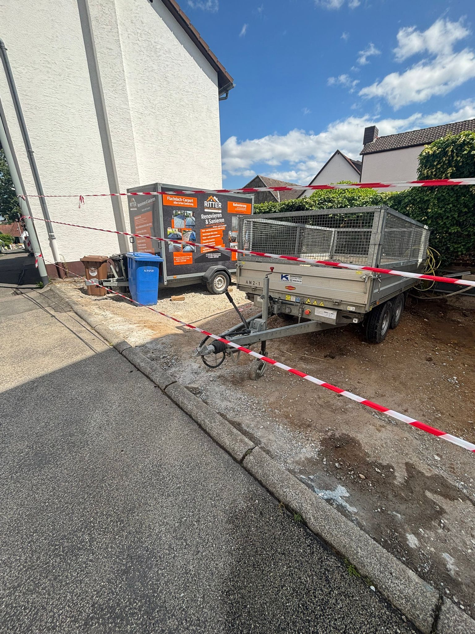 Ein Anhänger mit Metallzaunmaterialien steht neben einem Gebäude auf einer Schotterauffahrt; daneben befinden sich orangefarbene Bauschuttcontainer und Absperrband.
