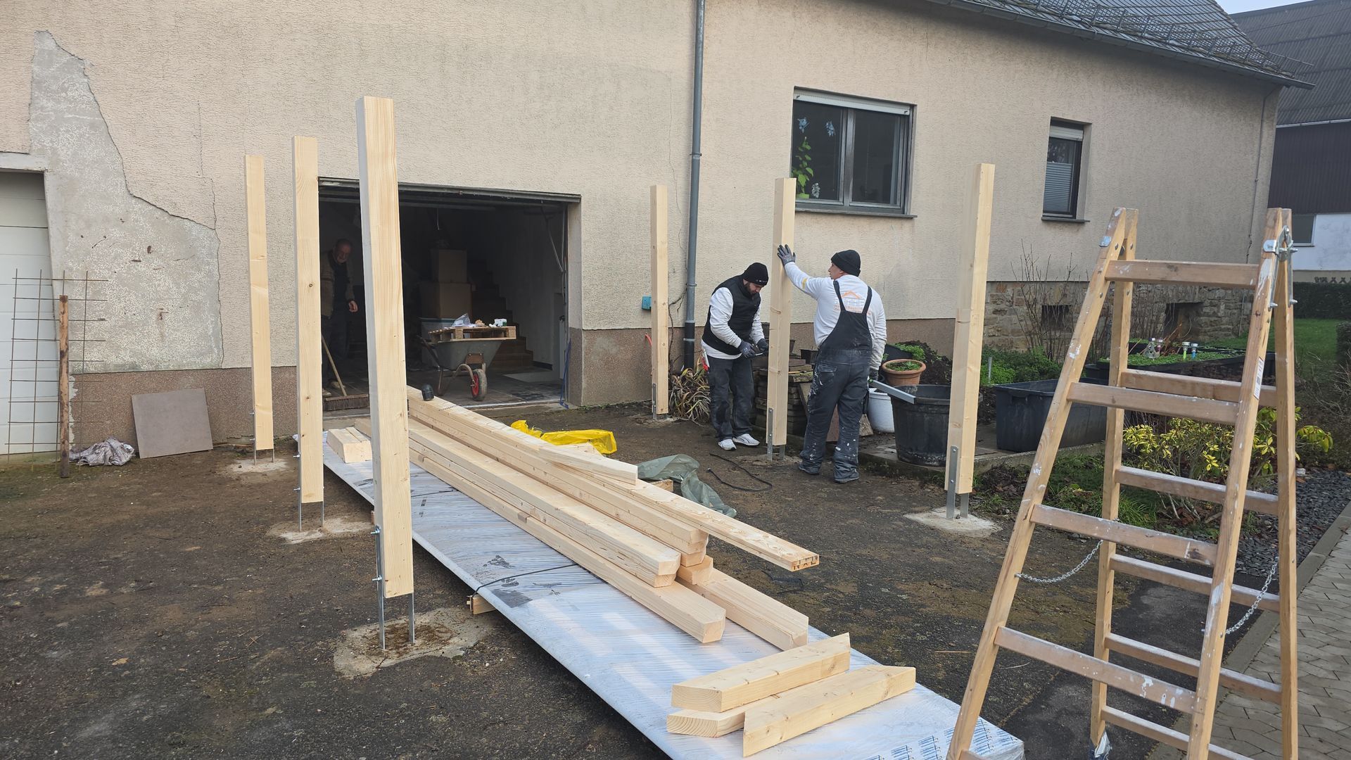 Errichtung einer Holzkonstruktion vor einer Garage. Zwei Personen arbeiten daran; Holz und eine Leiter sind vorhanden.
