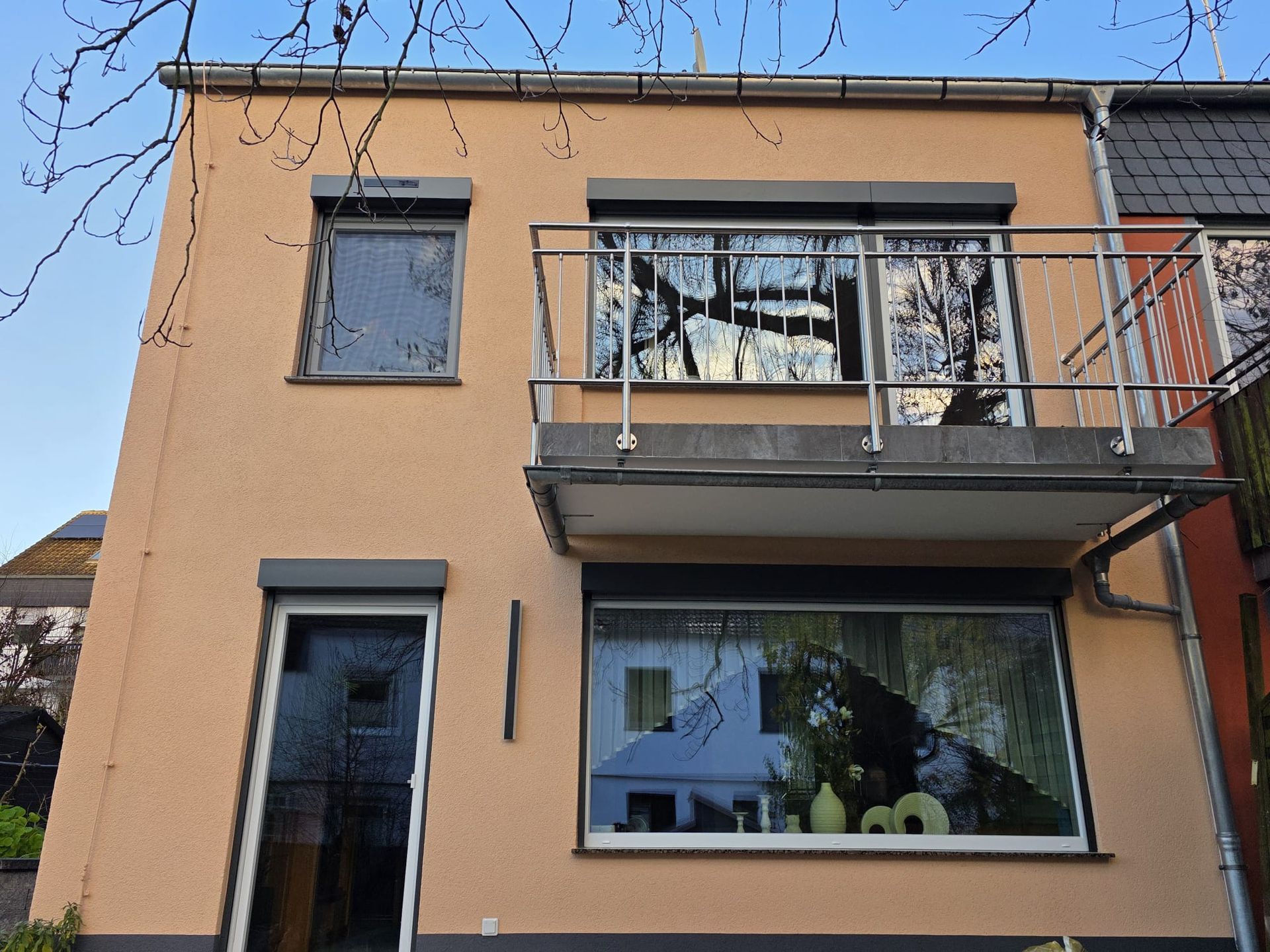 Beiges zweistöckiges Haus mit Balkon, großen Fenstern und Rollläden; blauer Himmel im Hintergrund.