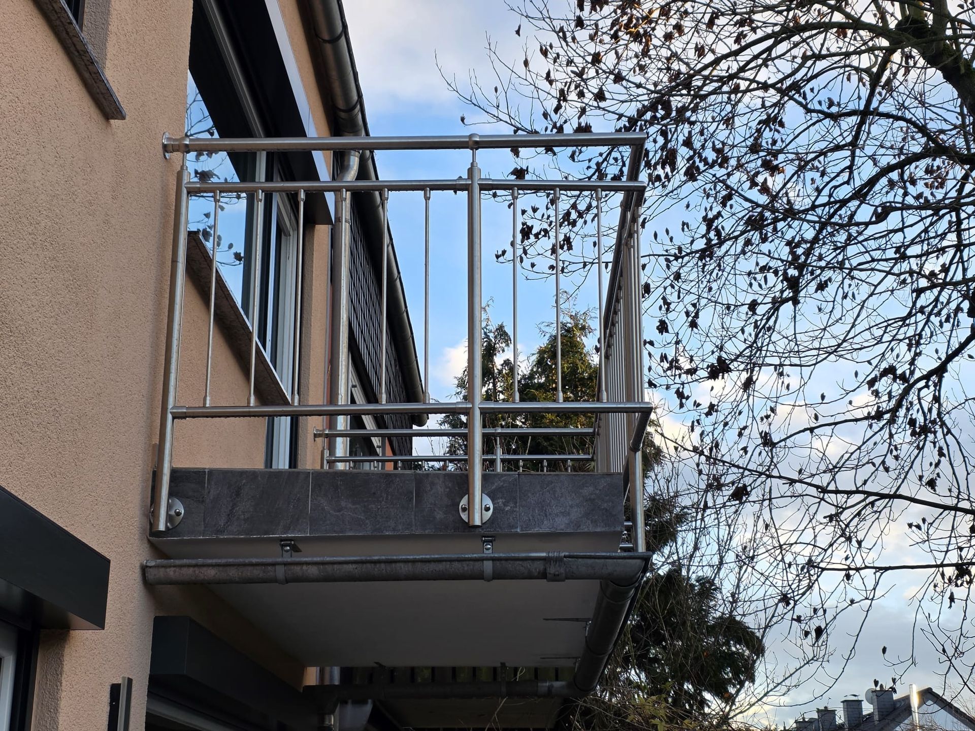Balkon mit Edelstahlgeländer an einem beigen Gebäude, im Hintergrund bewölkter Himmel und Baumzweige.