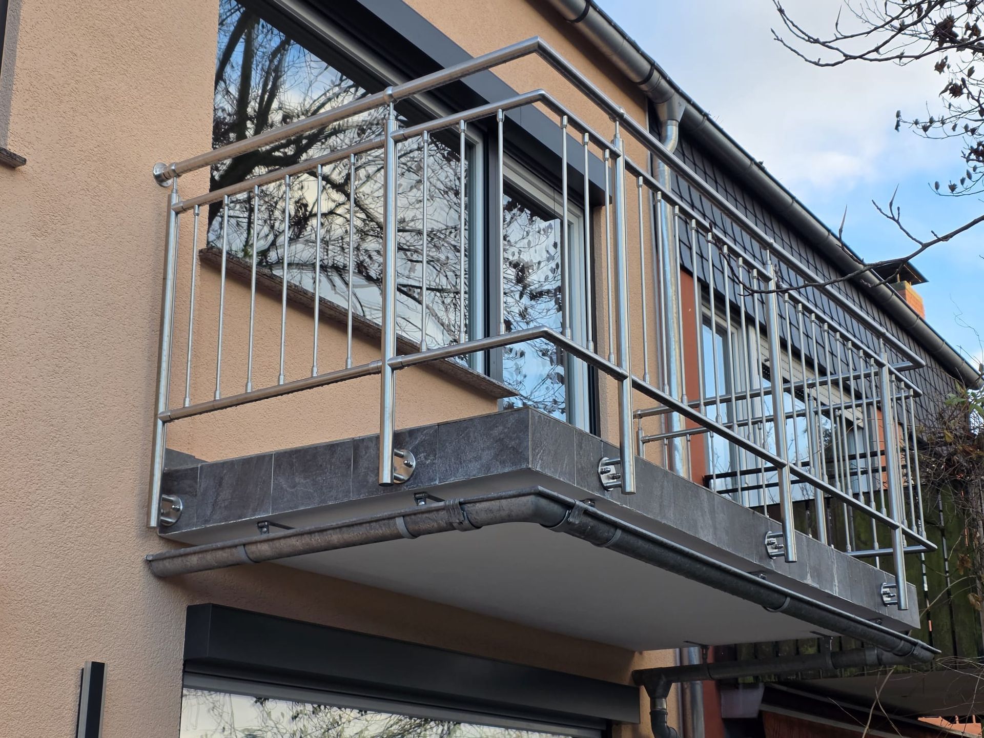 Balkon mit Edelstahlgeländer an einem beigen Gebäude mit Fenstern und Betonsockel.