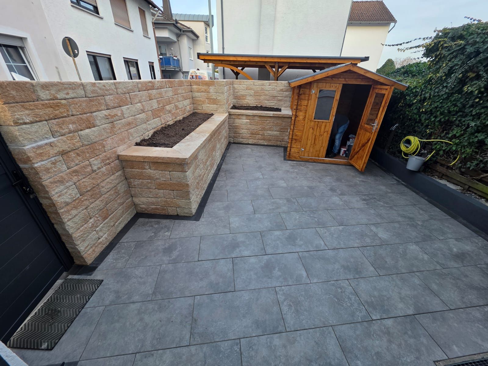 Mit Steinen gepflasterte Terrasse im Hinterhof mit erhöhten Sitzgelegenheiten aus Ziegelsteinen, einem Holzschuppen und umliegendem Grün