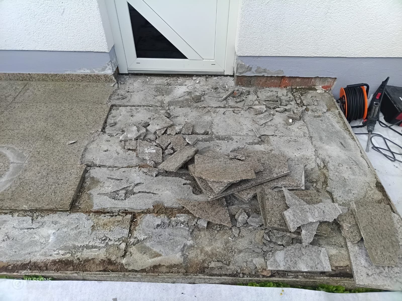 Vor einer weißen Tür befindet sich eine ausgebesserte Betonplatte, daneben liegen Bruchstücke von Trümmern und ein orangefarbener Werkzeugkasten.
