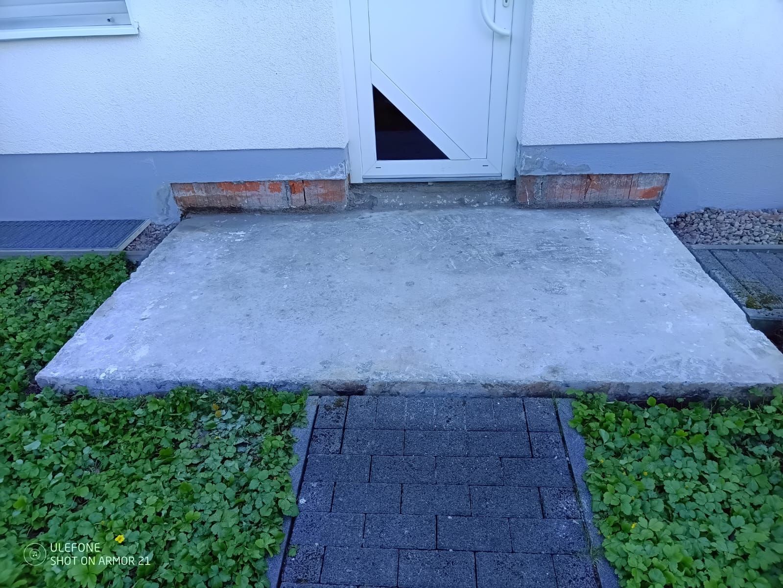 Eine barrierefreie Betonrampe führt zu einer weißen Tür mit einer kleinen Stufe und Gras auf beiden Seiten.