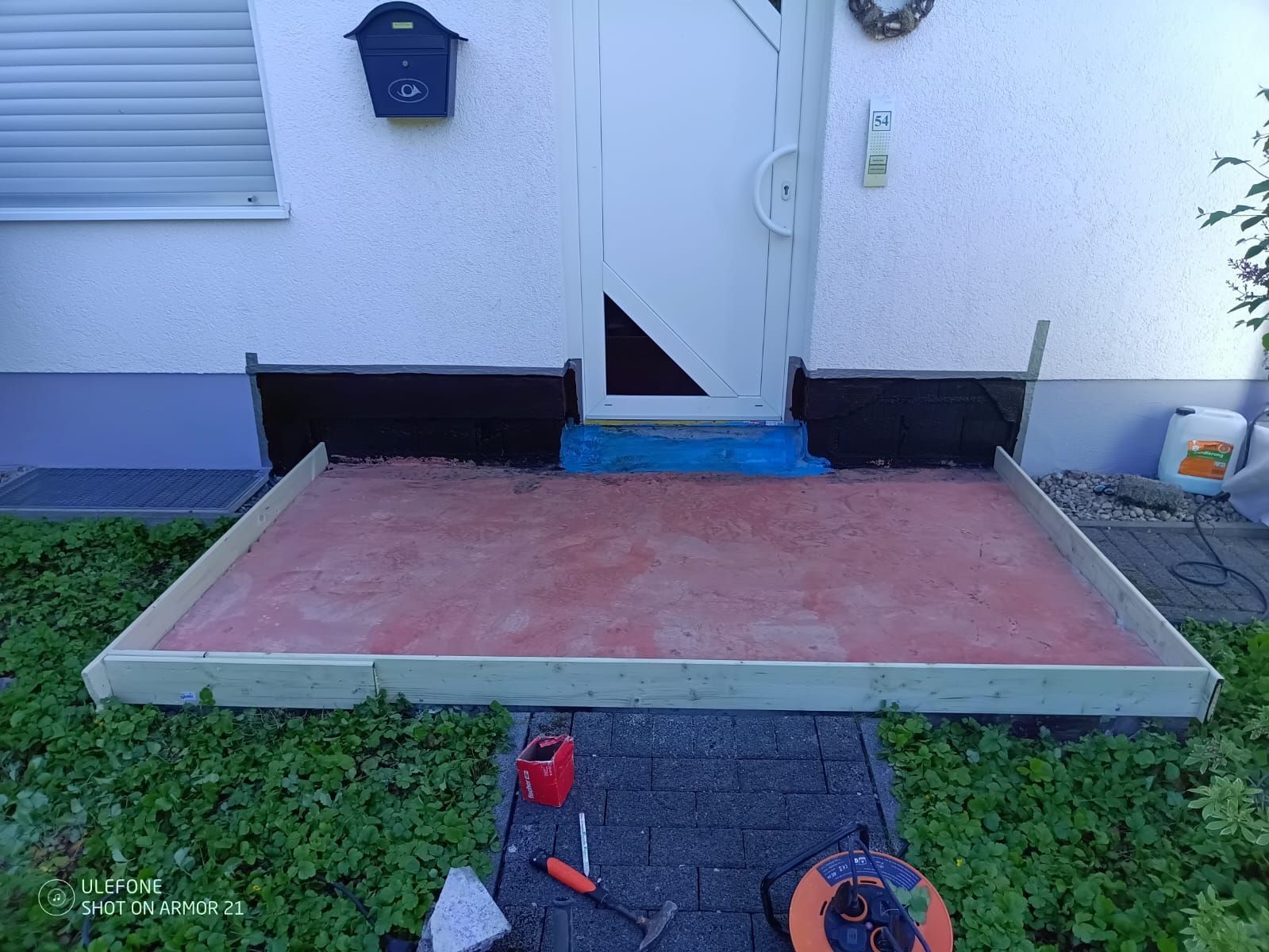 Eine kleine Betonplatte auf der Terrasse vor einer weißen Haustür, daneben Gartengeräte auf dem Rasen.