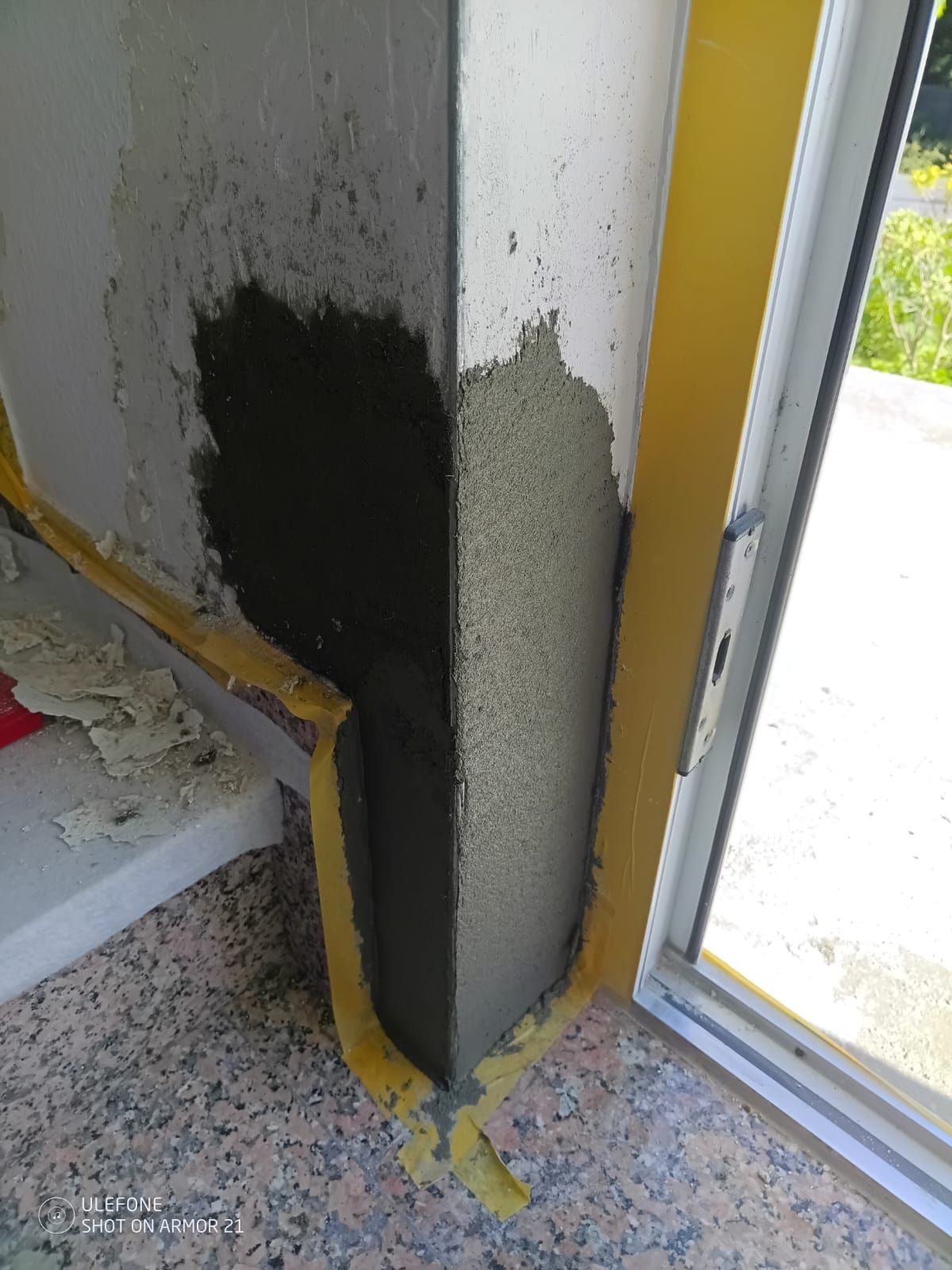 Eine teilweise verputzte Innenwandecke neben einer Tür, mit gelbem Klebeband und sichtbaren Ausbesserungsstellen.