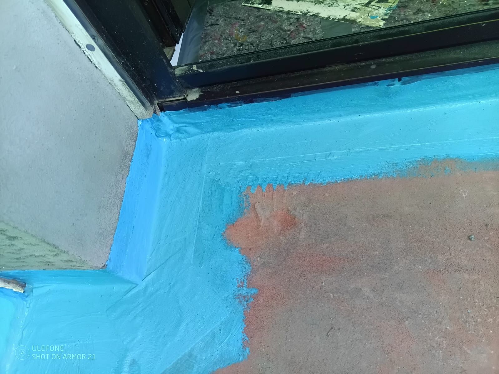 Eine blau gestrichene Ecke des Fußbodens neben einem Fenster, mit einem Stück blankem Beton.