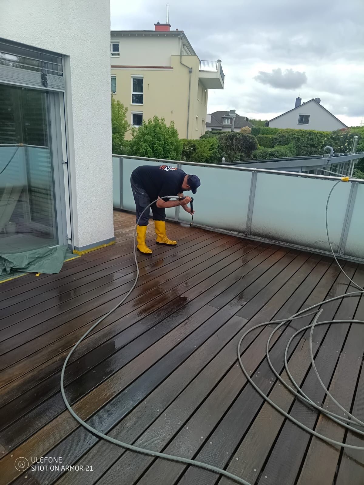 Eine Person reinigt mit einem Hochdruckreiniger eine nasse Holzterrasse neben einer Glastür; hinter dem Geländer sind Häuser zu sehen.