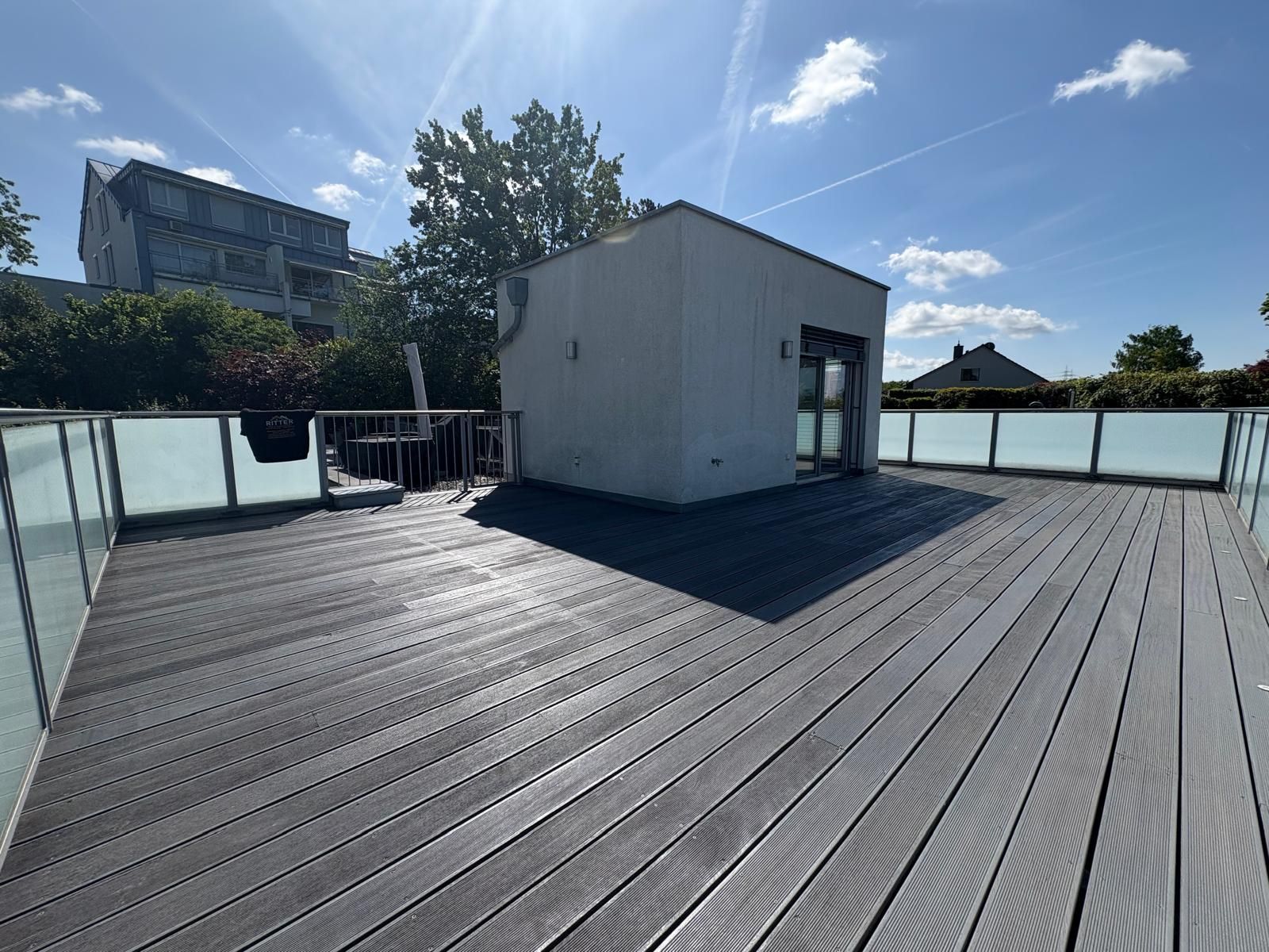 Dachterrasse mit grauen Holzplanken, Glasgeländer und einem kleinen weißen Geräteschuppen unter sonnigem, blauem Himmel