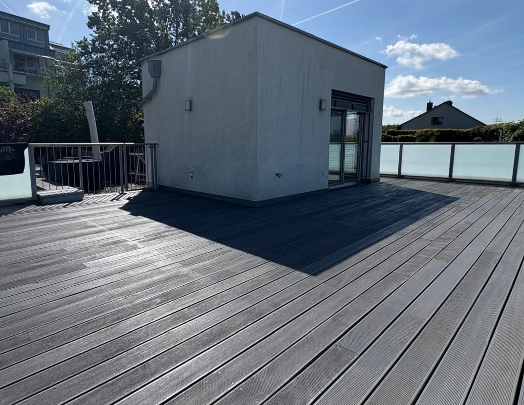 Dachterrasse mit dunklem Verbundboden und einem kleinen weißen Schuppen unter hellem Sonnenlicht
