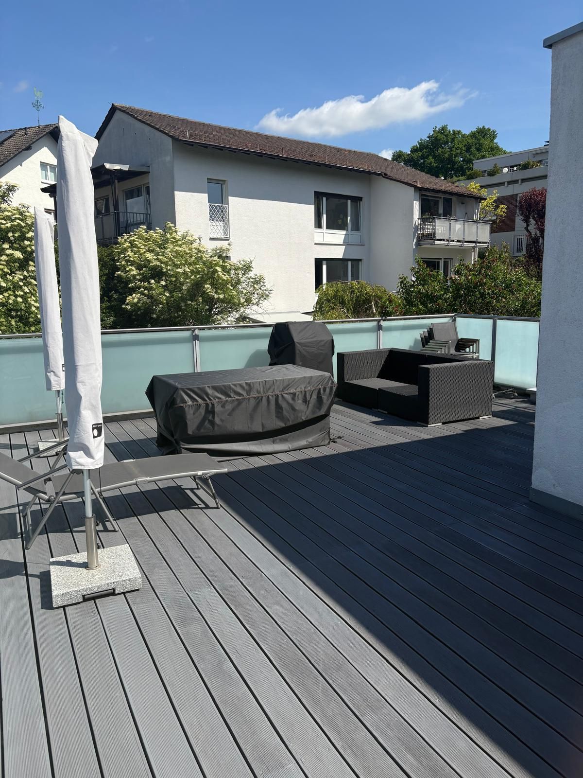 Dachterrasse mit grauen Sitzgelegenheiten, Glasgeländer und Blick auf benachbarte Wohnhäuser unter blauem Himmel