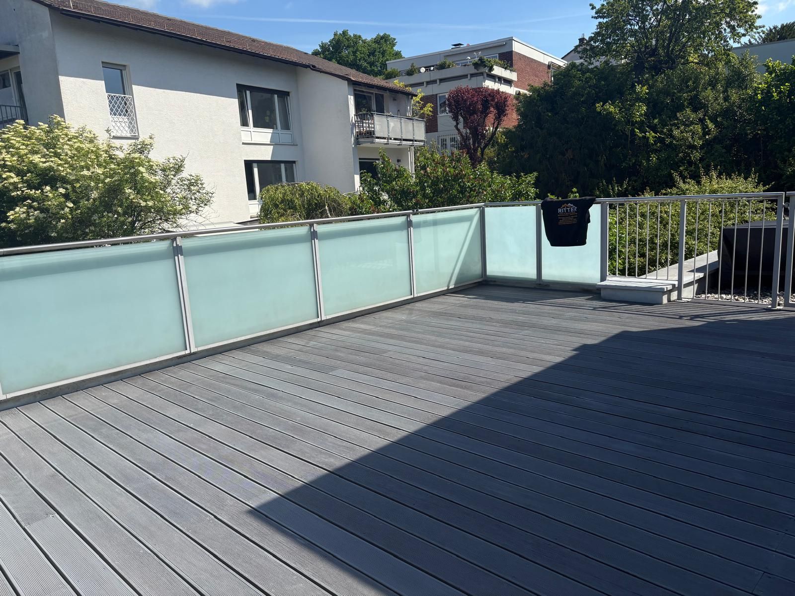 Holzterrasse mit Milchglasgeländer, Blick auf Häuser und Bäume an einem sonnigen Tag