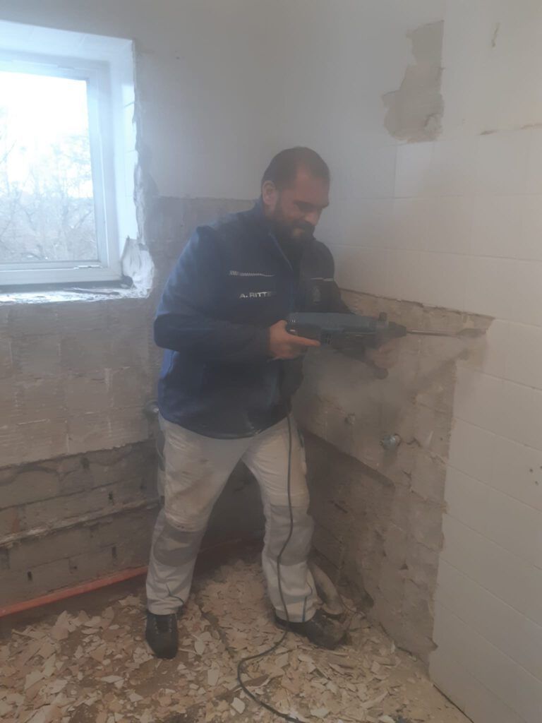 Ein Mann bohrt in einem Zimmer an einer Wand; die Wände sind beschädigt und staubig.