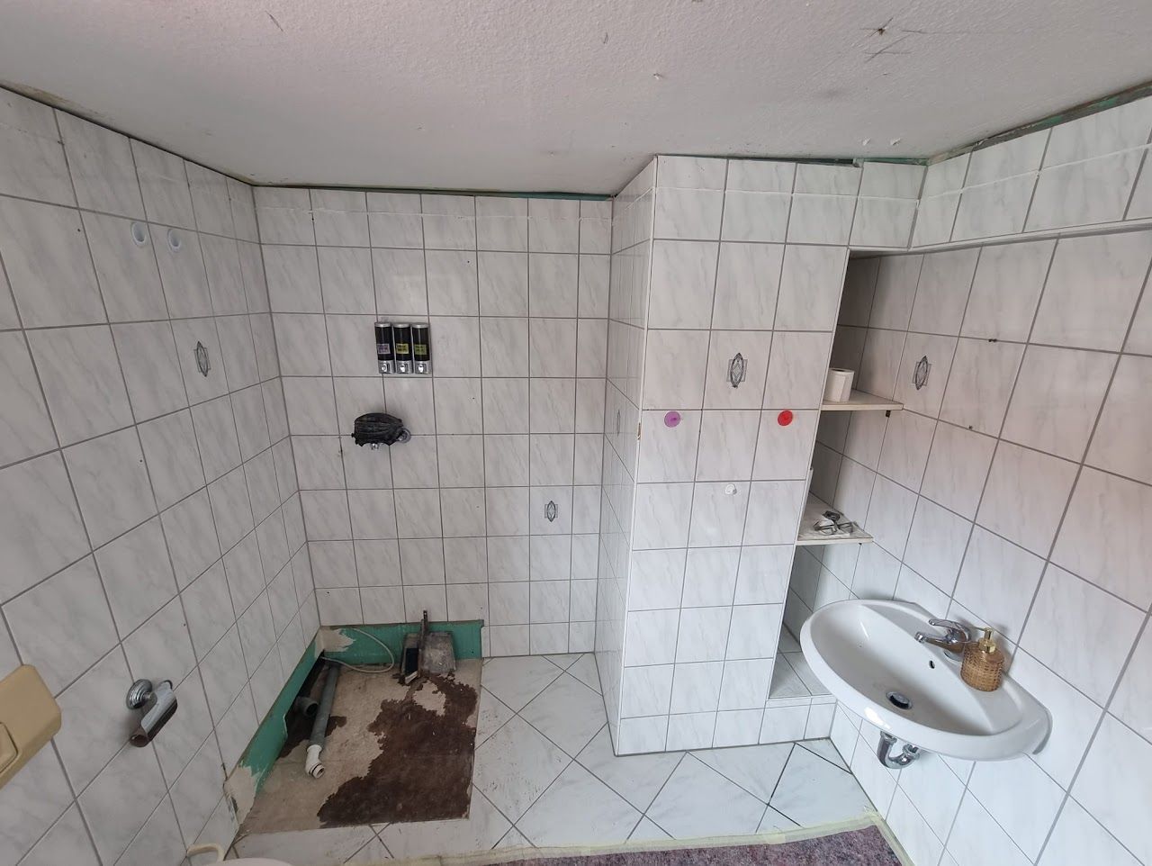 Badezimmer mit weiß gefliesten Wänden, freiliegenden Rohrleitungen und einem teilweise entfernten Bodenbelag.