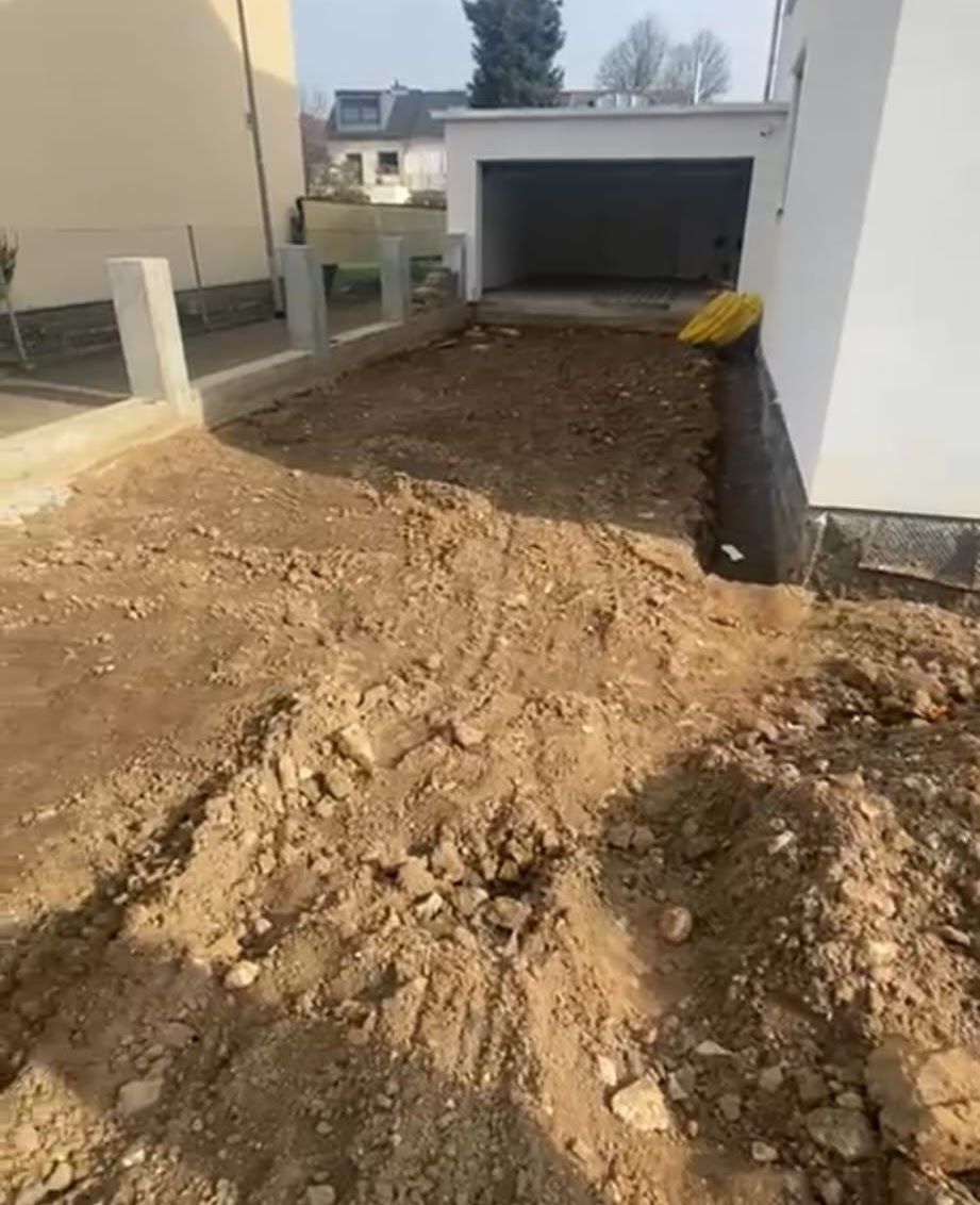 Unbefestigter Hof vor einer Garage, neben einer Betonmauer und einem Zaun.