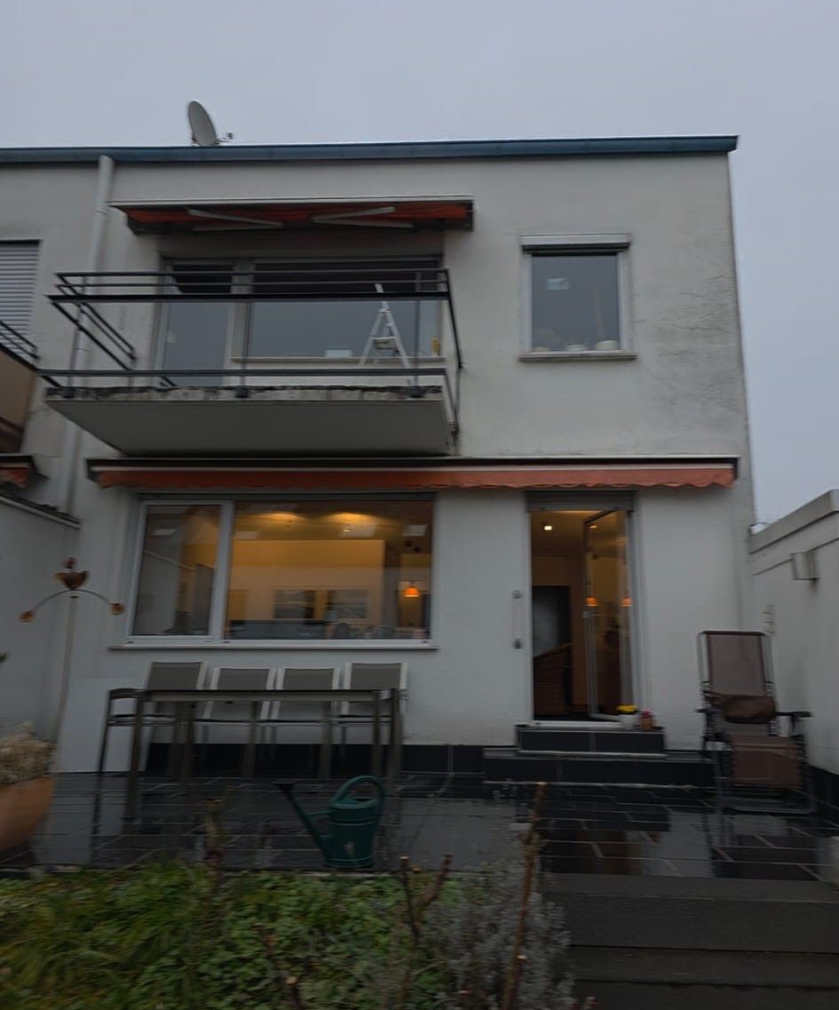Weißes zweistöckiges Haus mit Balkon, Markisen und offener Tür. Gartenmöbel und Treppe führen zum Eingang. Bewölkter Himmel.