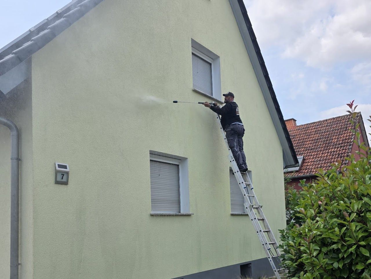 Ein Mann auf einer Leiter reinigt mit einem Hochdruckreiniger die Fassade eines hellen Hauses.