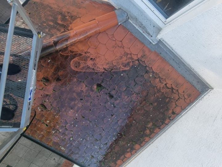 Draufsicht auf ein rotes Backsteindach mit Rissen und Moos, neben einer weißen Wand.