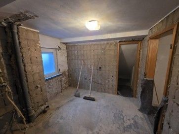 Zimmer im Umbau, freiliegende Betonwände, Fenster, offene Tür, Schutt auf dem Boden, zwei an die Wand gelehnte Besen.