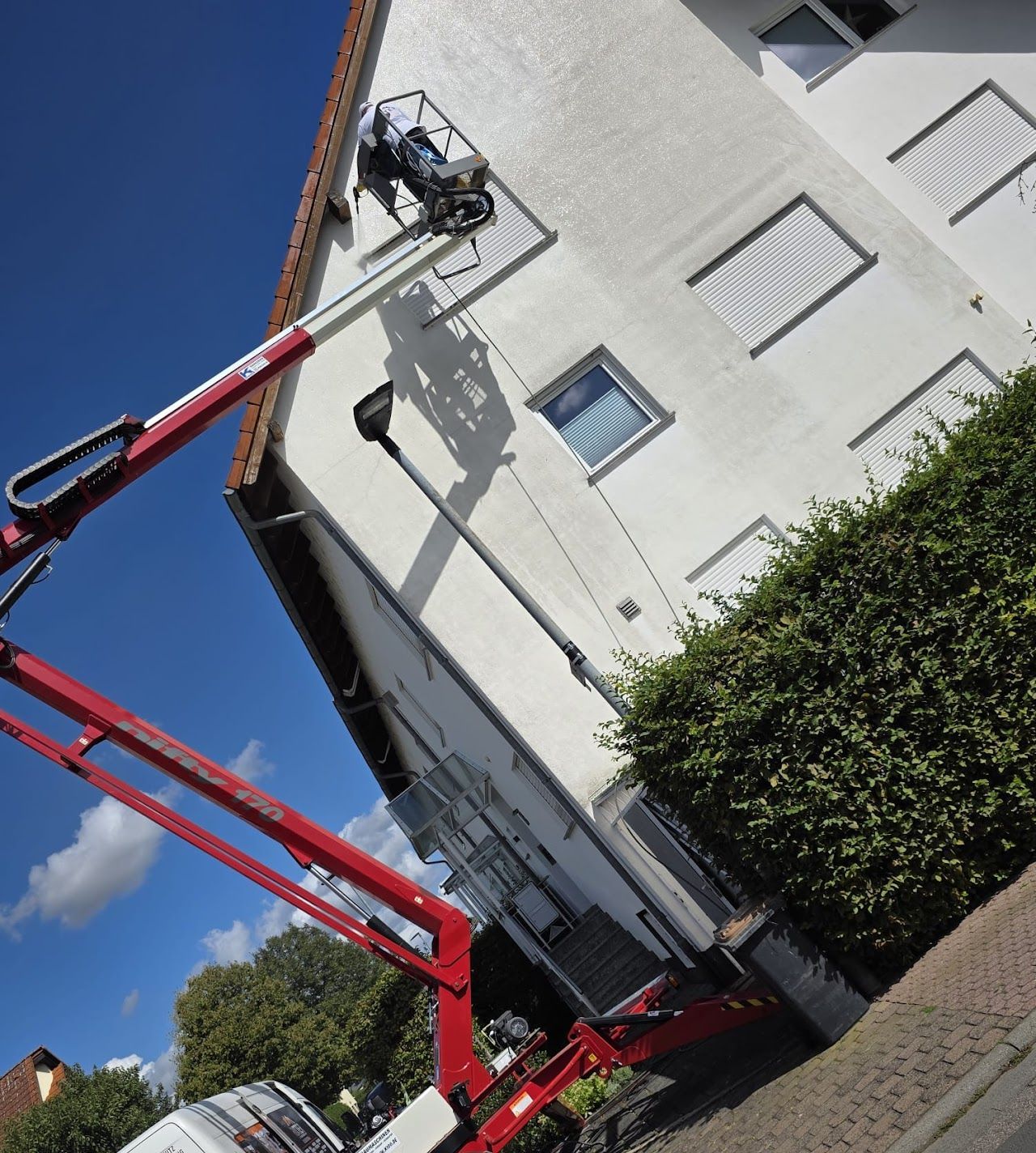 Ein roter Gabelstapler steht neben einem weißen Gebäude; eine Person arbeitet in einem Hubkorb an der Wand.
