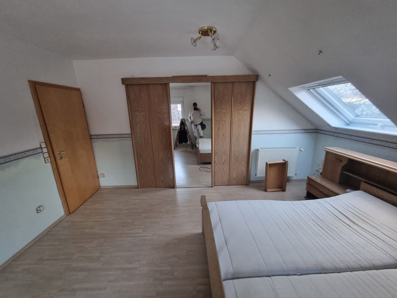 Schlafzimmer mit Bett, Schranktüren, Dachschräge und Dachfenster. Hellblaue Wand und Holzboden.