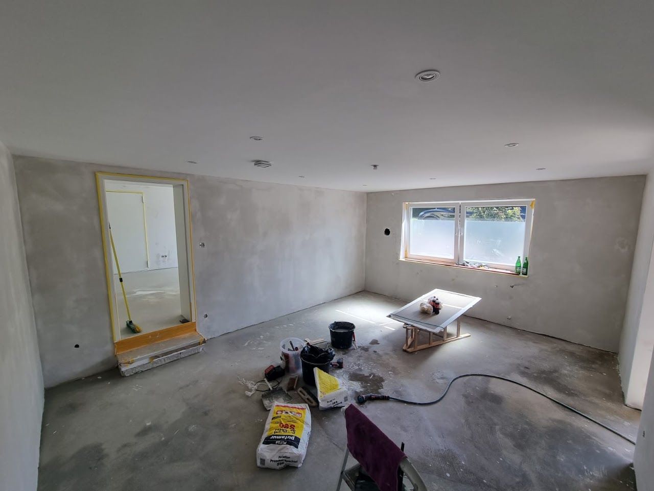 Ein Zimmer im Umbau; Wände verputzt, Betonboden unfertig. Tür und Fenster vorhanden, Werkzeuge und Materialien verstreut.