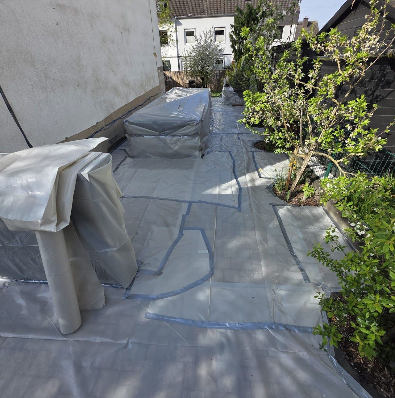 Die Terrasse ist mit Schutzfolie abgedeckt, die Möbel sind überdacht und an der Seite befindet sich Grün.