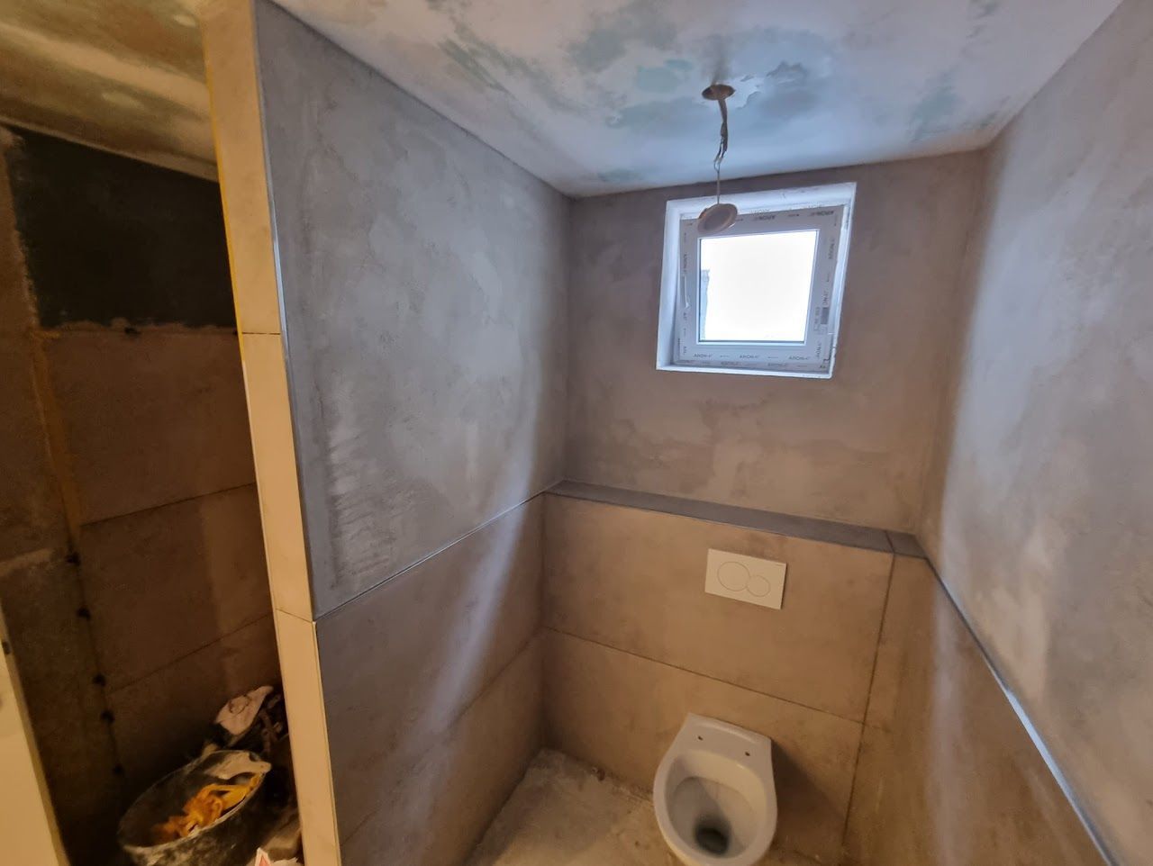 Kleines, unfertiges Badezimmer mit Toilette, Fenster und neuen Putzwänden.