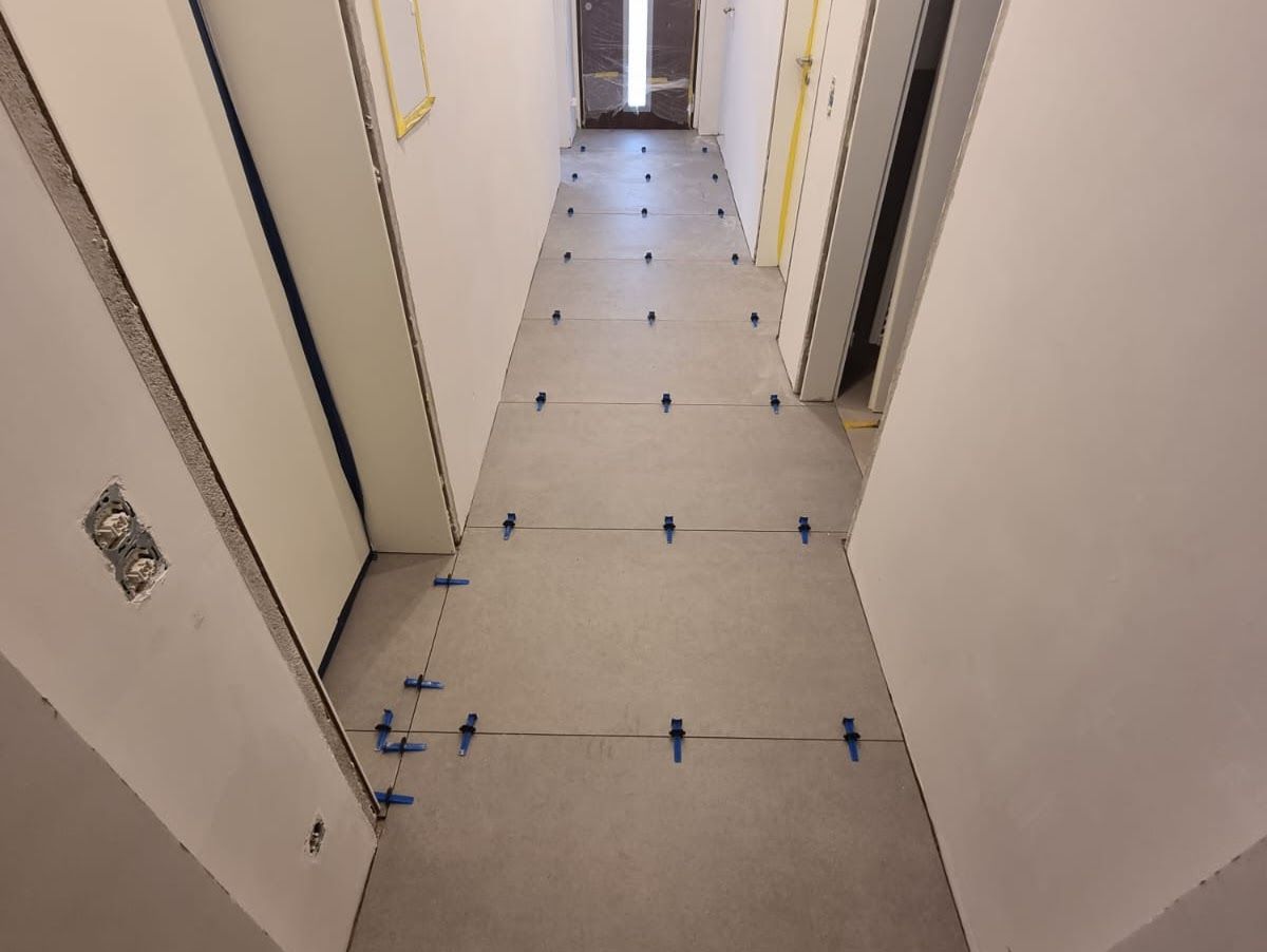 Flur, in dem gerade Bodenfliesen verlegt werden, Nivellierfüße zwischen den Fliesen sichtbar; unfertige Wände und Türrahmen.