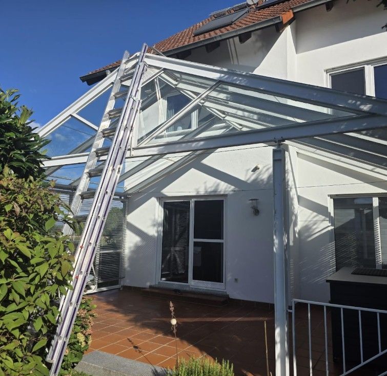 Eine Leiter lehnt an einer verglasten Terrasse, die an ein weißes Haus mit Terrakottafliesenboden angrenzt.