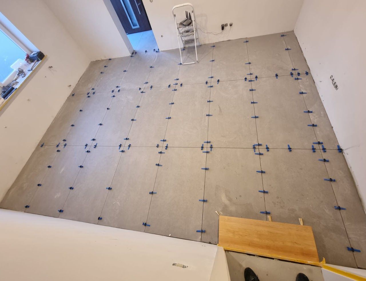 Es werden Bodenfliesen verlegt, grauer Boden mit blauen Fugenkreuzen und eine Leiter in einem Zimmer.