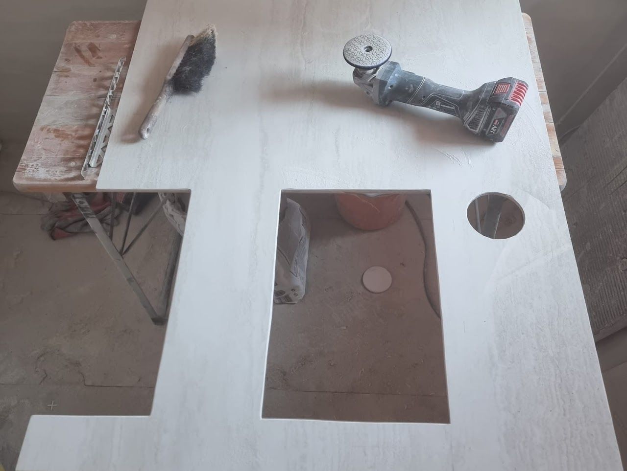 Eine helle Holzplatte mit ausgeschnittenen Abschnitten. Darauf befinden sich ein Winkelschleifer und eine Kelle.