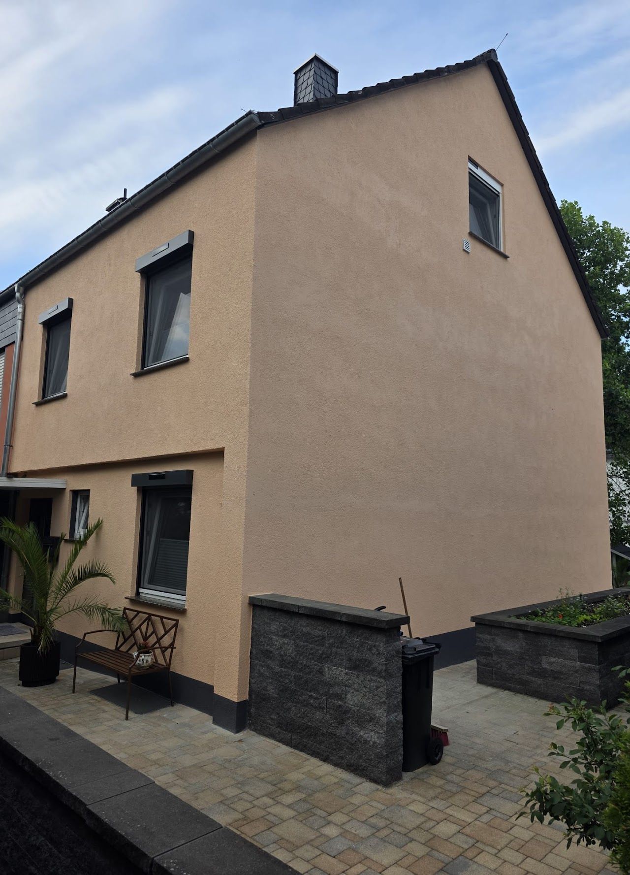 Beigefarbenes zweistöckiges Haus mit schwarzen Zierelementen, Fenstern und Dach; davor befindet sich eine Steinmauer.